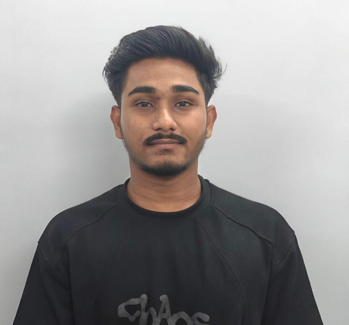 Young man with dark hair and beard wearing black T-shirt against plain background.